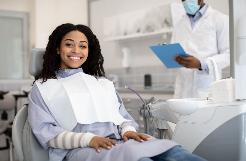 A woman seated in a dental chair and smiling while she wears a dental bib as her dentist stands behind her and examines a clipboard.