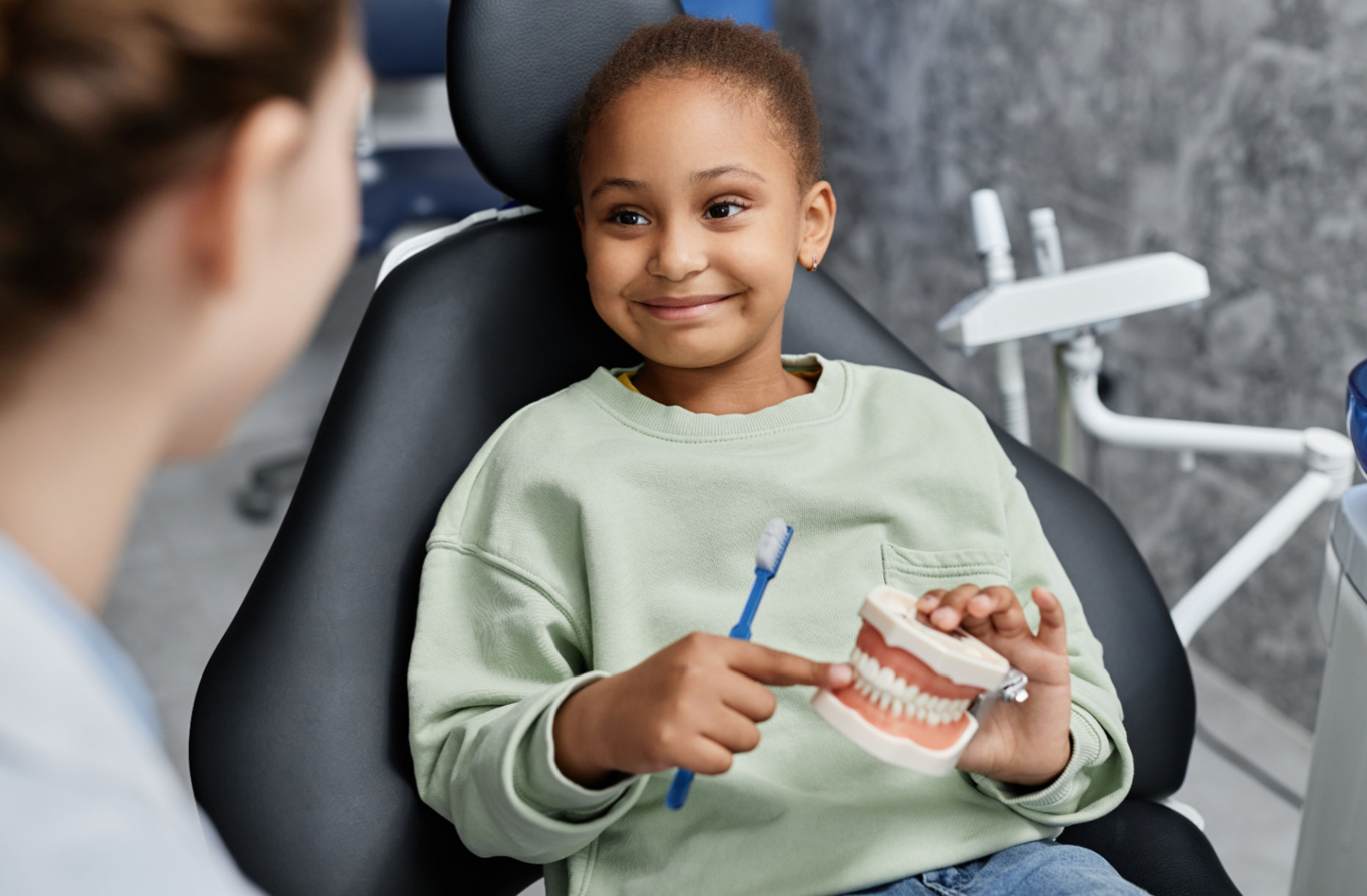 A smiling child in dental chair holding a model of teeth and a tooth brush.