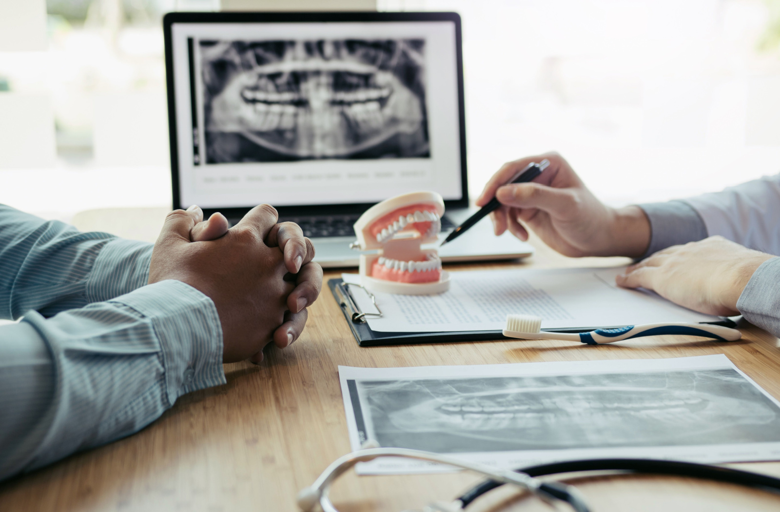 Two men's hands resting on a desk during a dental consultation. The dentist is using a pen to point to a dental model on the table, with a laptop showing an X-ray in the background.