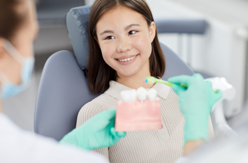 A young woman smiling in a dental chair as a dentist holds up a dental model and toothbrush in the foreground, out of focus.